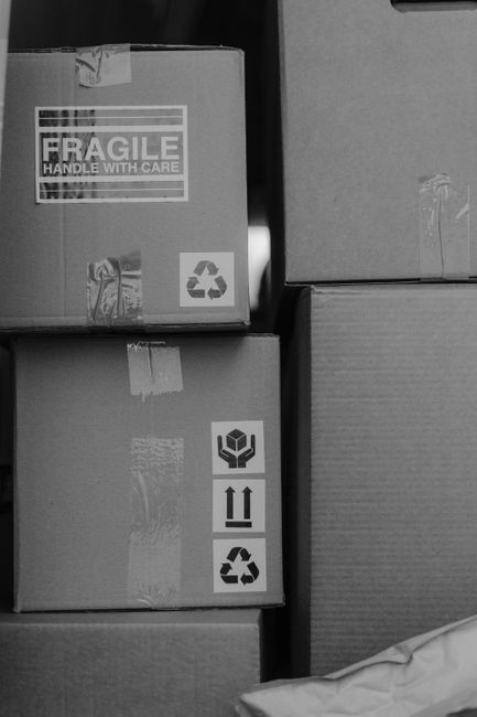 A close-up view of three stacked cardboard boxes in an indoor setting, with the top box displaying a large 'FRAGILE' label with handling instructions on a white background, including a recycle symbol 