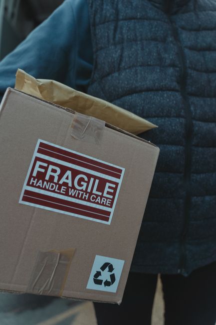 A close-up view of three stacked cardboard boxes in an indoor setting, with the top box displaying a large 'FRAGILE' label with handling instructions on a white background, including a recycle symbol 