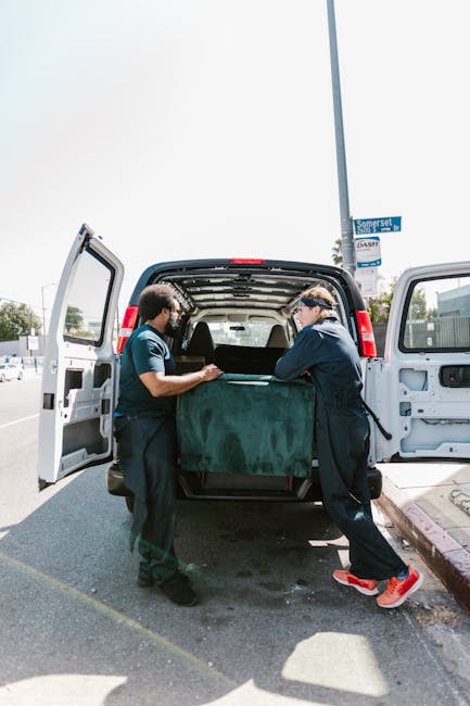 Two professional movers from Man and Van Vauxhall are engaged in loadings and furniture transport as part of a home relocation process. They are standing at the rear of a black van with its back doors open, inside which a large, green, padded moving blanket covers a piece of furniture or a bulky household item. Both movers are dressed in dark work uniforms and are carefully lifting or guiding the object into or out of the vehicle, indicating careful handling during packing and moving procedures. The van is parked on a paved street near a sidewalk, with clear visibility of a street sign and a lamppost indicating an urban environment. The background includes elements such as light posts and a few structures, suggesting a typical residential or commercial street. The scene demonstrates an organized loading process with the use of padding materials, relevant to the services offered by Man and Van Vauxhall in efficient and careful furniture transport within house removals and relocation services.