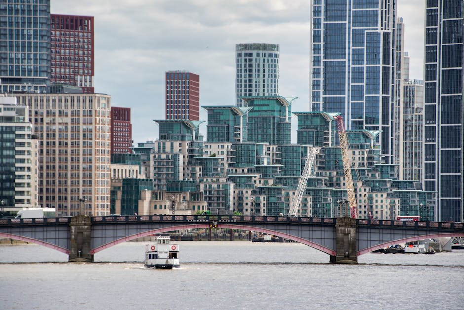 A cityscape view featuring modern high-rise buildings with glass facades and rooftop structures, situated behind a river with a small boat passing underneath a stone arch bridge. Two cranes are visible among the buildings, indicating ongoing construction or development. The scene is captured during daylight with a partly cloudy sky, and the river reflects the urban environment. The image highlights urban architecture, construction activity, and city logistics, aligning with themes of property relocation and moving services offered by Man and Van Vauxhall, as seen on their website, manandvanvauxhall.co.uk.