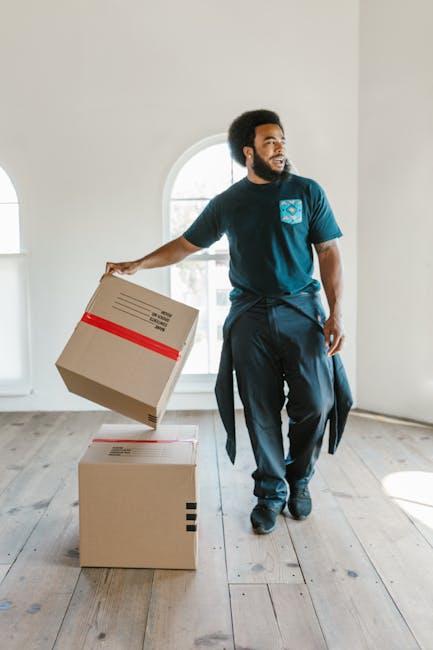 Two professional movers from Man and Van Vauxhall are engaged in loadings and furniture transport as part of a home relocation process. They are standing at the rear of a black van with its back doors open, inside which a large, green, padded moving blanket covers a piece of furniture or a bulky household item. Both movers are dressed in dark work uniforms and are carefully lifting or guiding the object into or out of the vehicle, indicating careful handling during packing and moving procedures. The van is parked on a paved street near a sidewalk, with clear visibility of a street sign and a lamppost indicating an urban environment. The background includes elements such as light posts and a few structures, suggesting a typical residential or commercial street. The scene demonstrates an organized loading process with the use of padding materials, relevant to the services offered by Man and Van Vauxhall in efficient and careful furniture transport within house removals and relocation services.