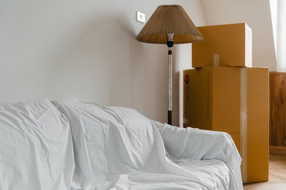 In a room with natural daylight coming from a window, there are three cardboard moving boxes stacked in a corner near the wooden furniture. A floor-standing lamp with a brown fabric shade is positioned next to the boxes, casting a soft light on the space. In the foreground, a white fabric-covered sofa with slightly wrinkled upholstery is partially visible. The setting appears inside a home during a packing or home relocation process, with items prepared for a move by [COMPANY_NAME], highlighting the typical scene of furniture and boxes in a residence awaiting transport.