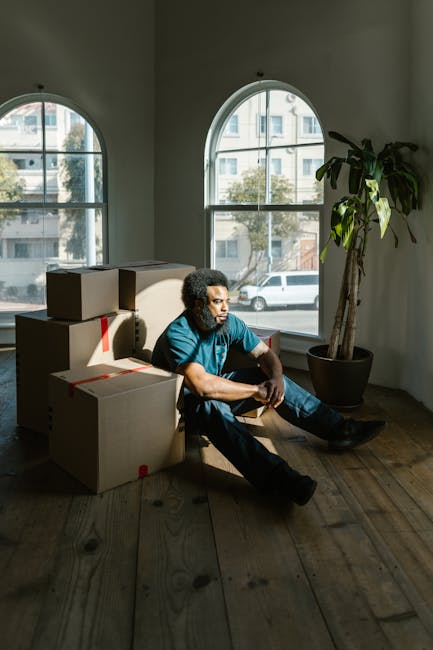 A man with curly hair, wearing a teal T-shirt and jeans, is seated on a wooden floor inside a room with large arched windows. Next to him are several cardboard boxes, some sealed with red tape, indicating packing for a home relocation or moving process. The room is illuminated by natural daylight coming through the windows, which reveal a city street with parked cars and residential buildings. A tall potted plant with broad leaves is positioned near the windows, adding greenery to the space. The scene suggests the initial stages of furniture transport or moving preparations, with the man possibly taking a break during packing or loading activities. This image exemplifies the furniture and packing materials involved in professional removals, such as those offered by Man and Van Vauxhall, supporting a comprehensive house move or delivery service.