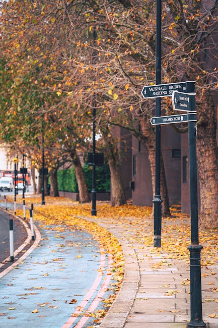In a room with natural daylight coming from a window, there are three cardboard moving boxes stacked in a corner near the wooden furniture. A floor-standing lamp with a brown fabric shade is positioned next to the boxes, casting a soft light on the space. In the foreground, a white fabric-covered sofa with slightly wrinkled upholstery is partially visible. The setting appears inside a home during a packing or home relocation process, with items prepared for a move by [COMPANY_NAME], highlighting the typical scene of furniture and boxes in a residence awaiting transport.