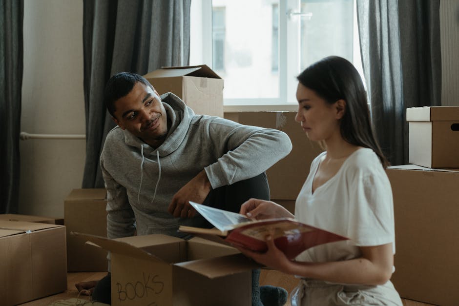 Inside a residential room during a home relocation, a young man with short dark hair, wearing a light grey hoodie, is sitting on the floor amid stacked cardboard boxes, some of which are labeled with 